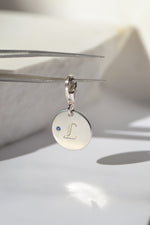A solid sterling silver pendant, engraved with pet's 'L' initial, accented by a natural round brilliant-cut blue sapphire. REINE by PAWVÉ.