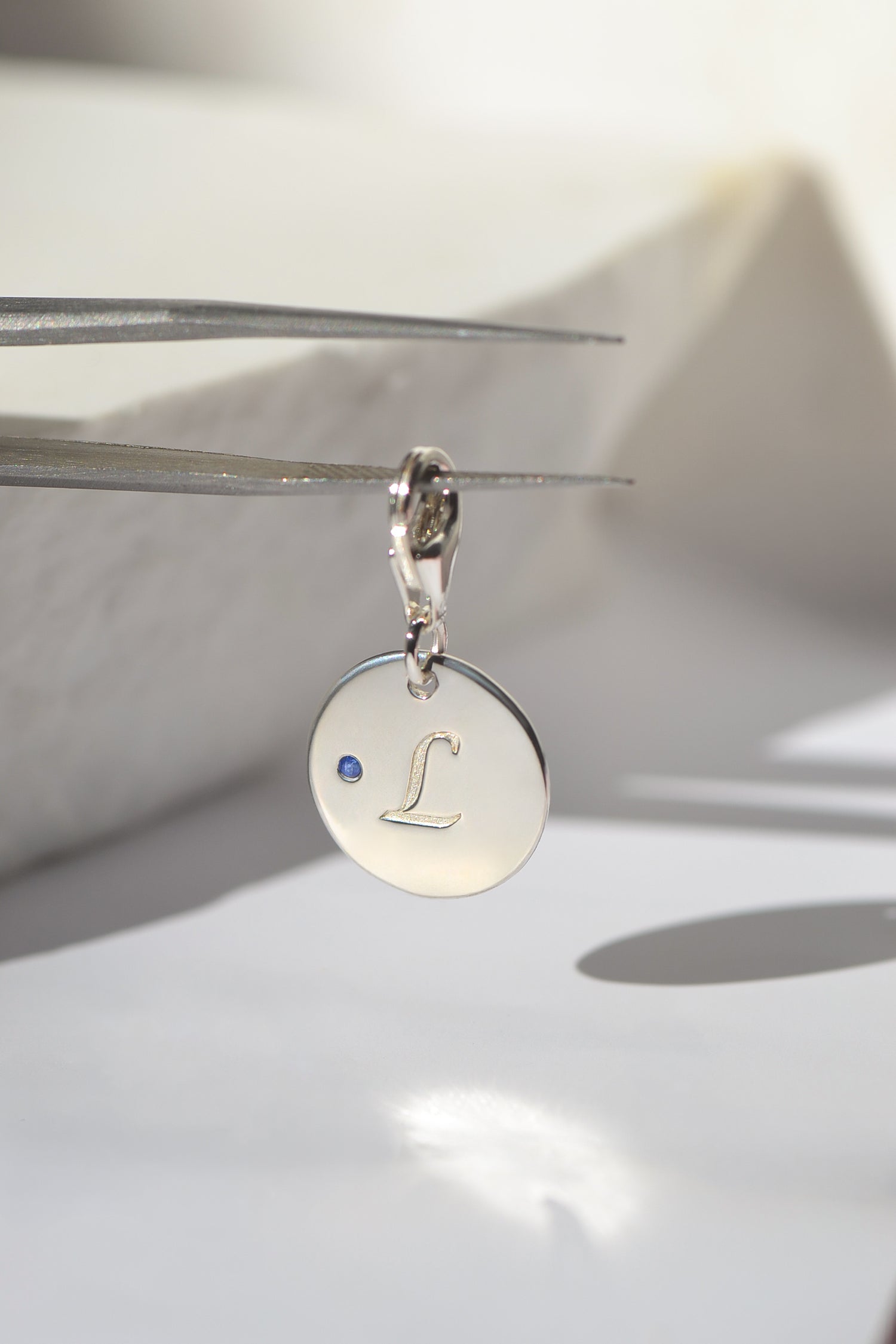 A solid sterling silver pendant, engraved with pet's 'L' initial, accented by a natural round brilliant-cut blue sapphire. REINE by PAWVÉ.